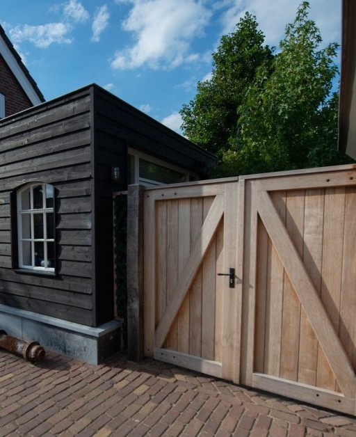 Een robuuste, dubbele houten opritpoort van onbehandeld eikenhout naast een bijgebouw met zwarte houten gevelbekleding. De poort heeft een klassiek Z-frame en zwart beslag, geplaatst op een bestrating van authentieke waaltjes onder een blauwe lucht.